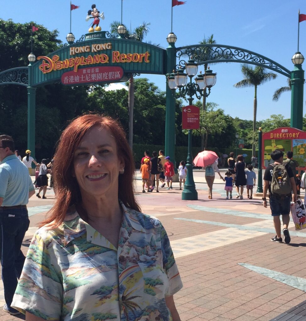 Kathryn Woodcock (white woman, auburn hair, (seen only from elbows up) poses in front of entrance sign of Hong Kong Disneyland Resort, on a sunny day, blue sky, palm trees, not too crowded; wearing aloha shirt