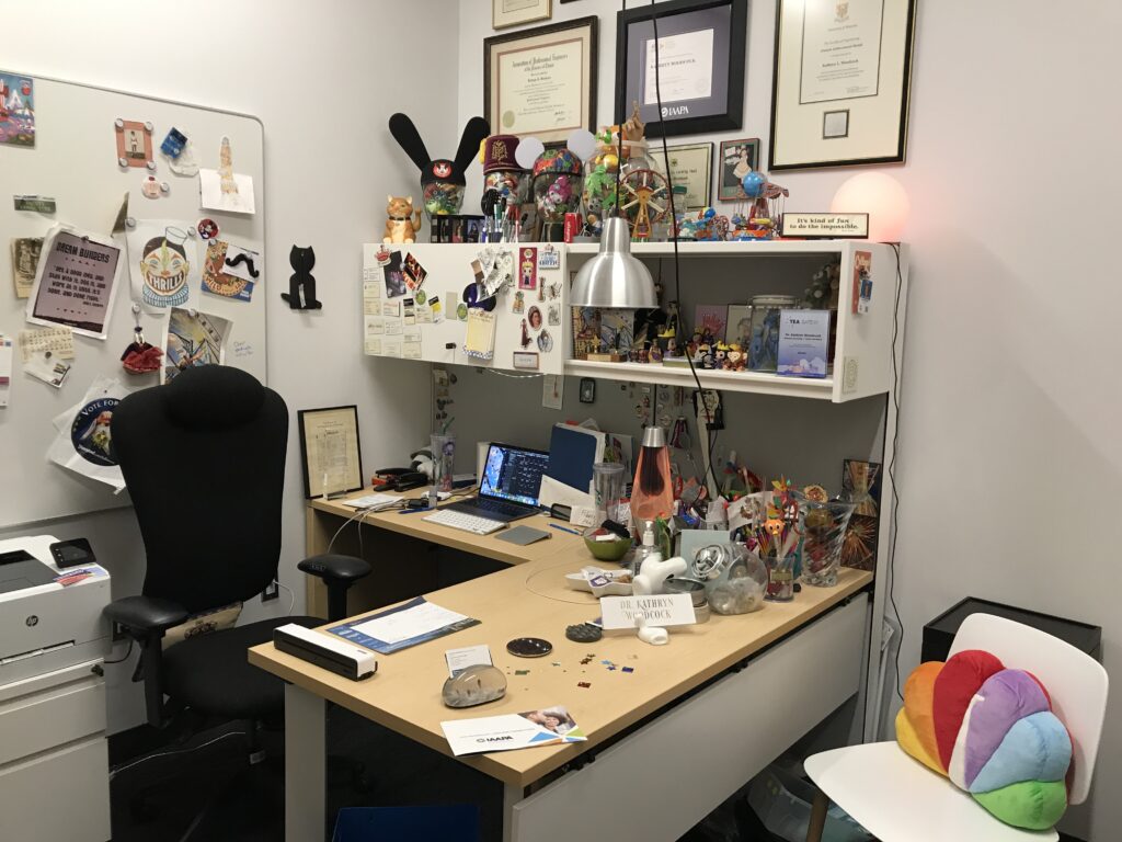 ivory office desk with blonde wood top, ivory wall-mounted overhead bin, with lots of toys and a lava lamp; a white chair in the foreground has an NBC peacock throw pillow