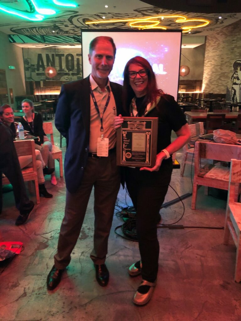 Steve Blum (white man in suit and name tag lanyard) presents plaque of US patent to Kathryn Woodcock (white woman, wearing glasses, black top and pants) in dining room at Antijitos Universal CItyWalk
