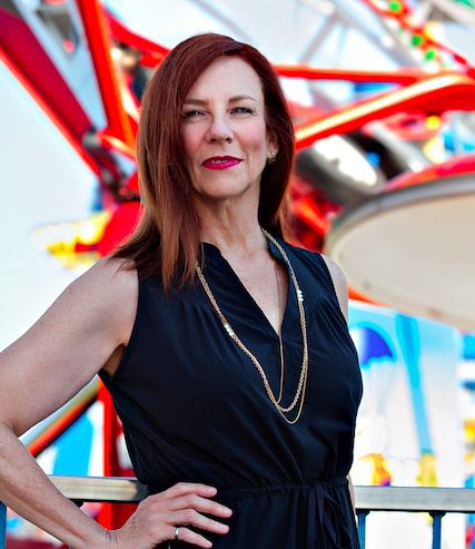 Photo courtesy of TSSA, from Safety Impact Award profile: Kathryn Woodcock, white woman, auburn hair, in sleeveless black dress, gold chain poses in front of roller coaster at Fun Spot, Orlando
