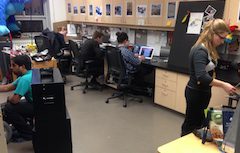 THRILL Lab workspace, blonde wood cabinets, black desktop, pictures taped on cupboard doors; several diverse students work on various things in the space.