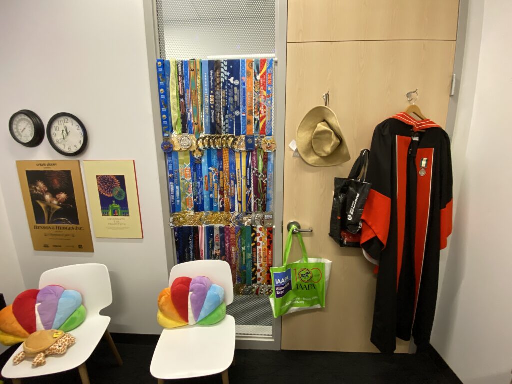 View of office door from inside: visible UofT doctoral robe with medal on facing, sun hat, two white chairs both with NBC peacock throw cushions, two Symphony of Fire posters on wall, below clock and thermometer, and dozens of half marathon medals hung up as window covering beside door