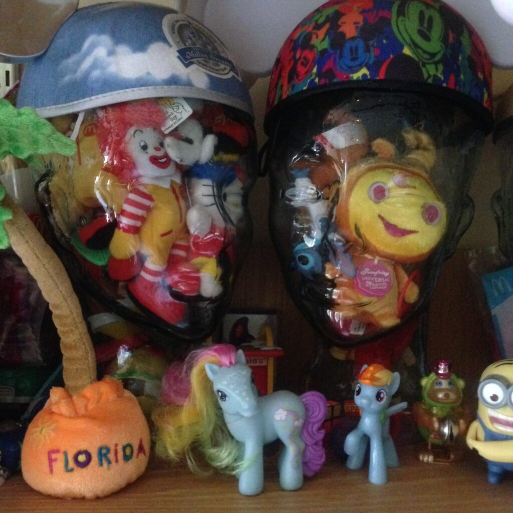 Two glass wig stands wearing Disney mouse ears stuffed with a variety of happy-meal toys and souvenir plush items, with some "my little pony" etc. in foreground - purely whimsical decorative image