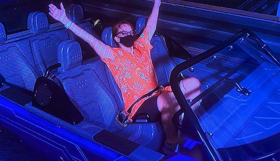 blurry photo of monitor picture showing Kathryn Woodcock seated alone in 6-seat ride vehicle at Test Track, wearing gaudy orange aloha shirt, black shorts, and black COVID mask