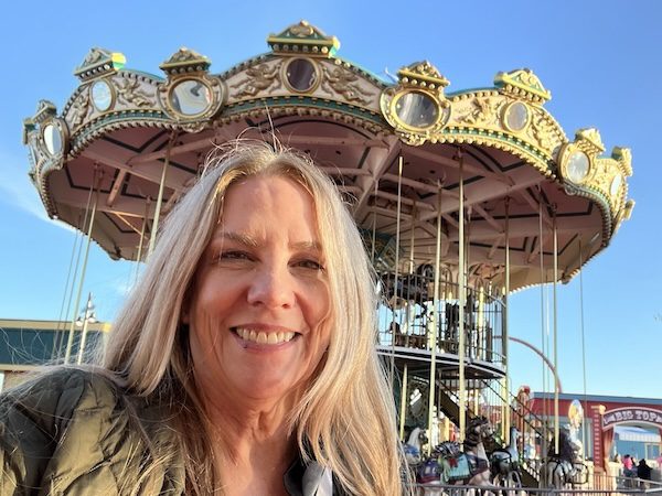 KW head in front of two-tier carousel. Blonde hair, smiling. Carousel has gold trim. Blue sky beyond.