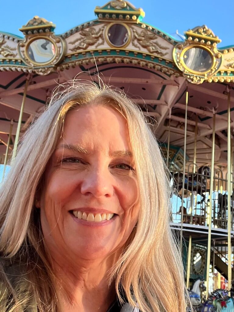 KW head in front of carousel. Blonde hair, smiling. Carousel has gold trim. Blue sky beyond.