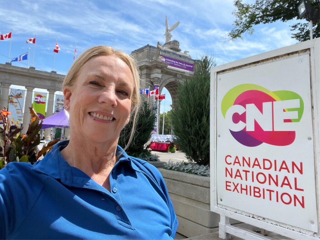 Kathryn Woodcock (white woman, hair in ponytail) selfie in front of Princes' Gates with sign for CNE Canadian National Exhibition