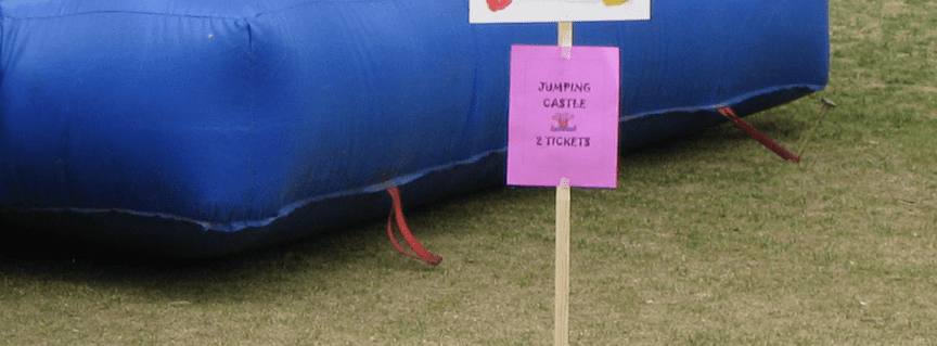 close up of base of inflatable with one tether anchored to a bare stake protruding threateningly almost a foot above the ground, and the other tether not staked at all. In the foreground, a sign indicates "Jumping castle 2 tickets"